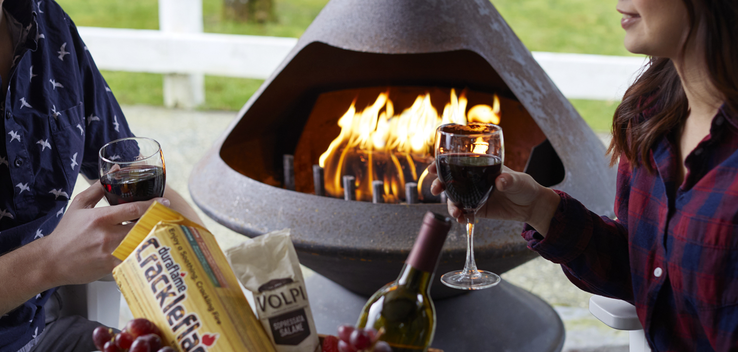 Two people drinking wine together and talking with a Duraflame Crackleflame log burning in an outdoor fireplace in the background