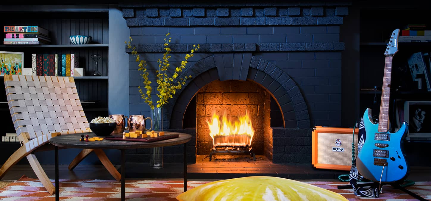 Cozy living room with a black brick fireplace and warm fire. Stylish decor includes a blue electric guitar, low wooden chair, and table with drinks, perfect for relaxing.