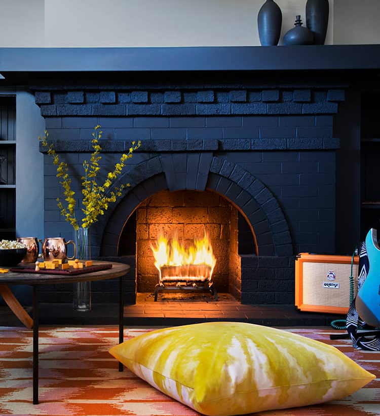 Cozy living room with a black brick fireplace and warm fire. Stylish decor includes a blue electric guitar, low wooden chair, and table with drinks, perfect for relaxing.