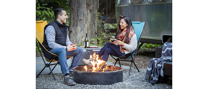 Glamping with Airstream in background and couple enjoying DURAFLAME® OUTDOOR FIRELOGS burning in campfire pit