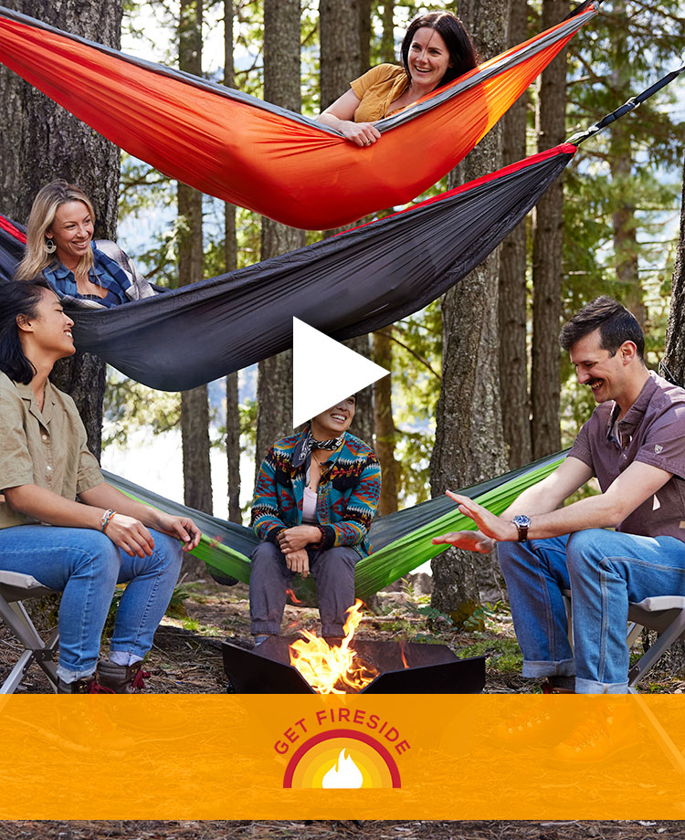 Group of people in forest in hammocks and chairs sitting by an outdoor fire with art with flame that says 